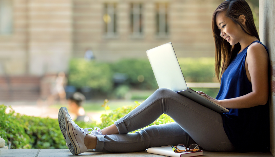 A female Asian learner sits outside on a college campus on a sunny day with her back against a wall and her legs stretched out on a ledge studying on her laptop