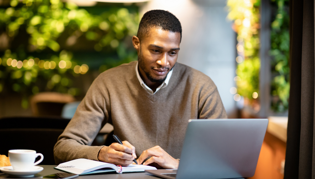 A man sits at a table, writing in a notebook while looking at his laptop.