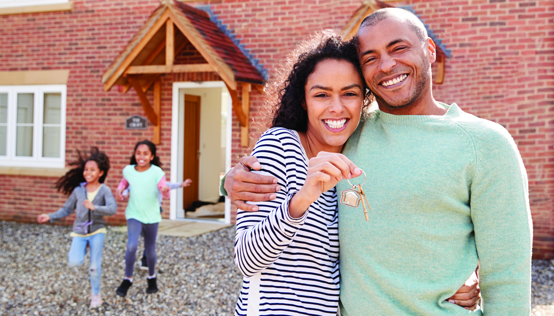 Smiling couple holding keys to new home with children playing in the background