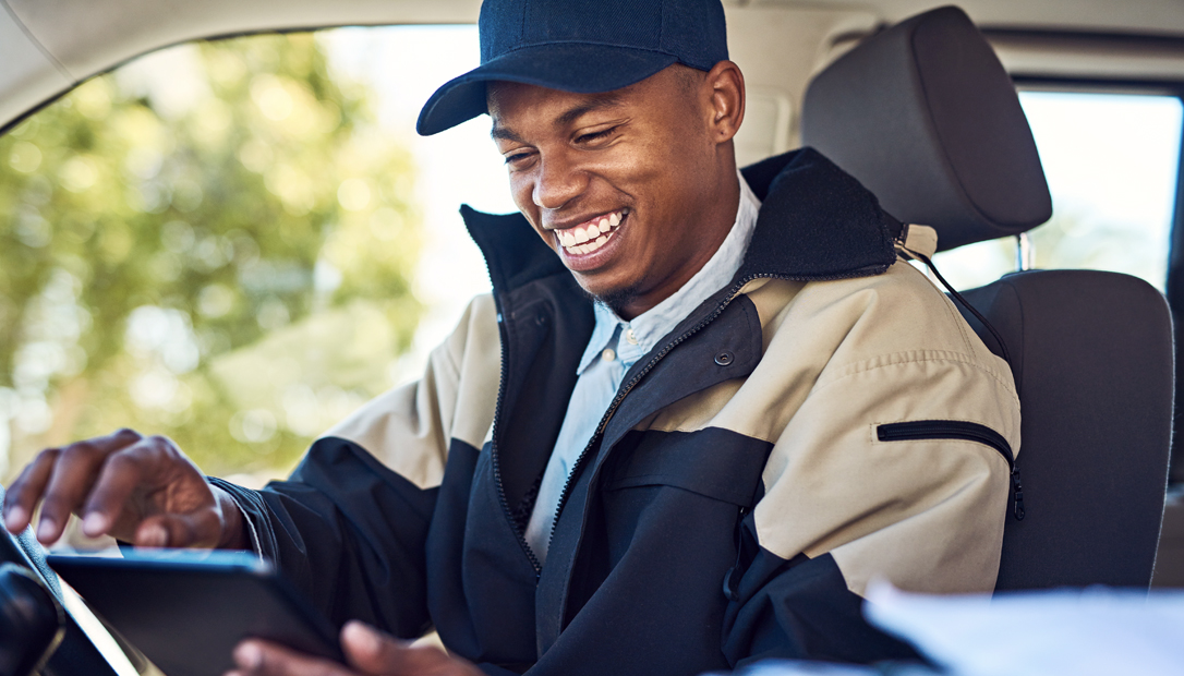 A Black Amazon delivery driver sits in his vehicle while smiling and working on a tablet 