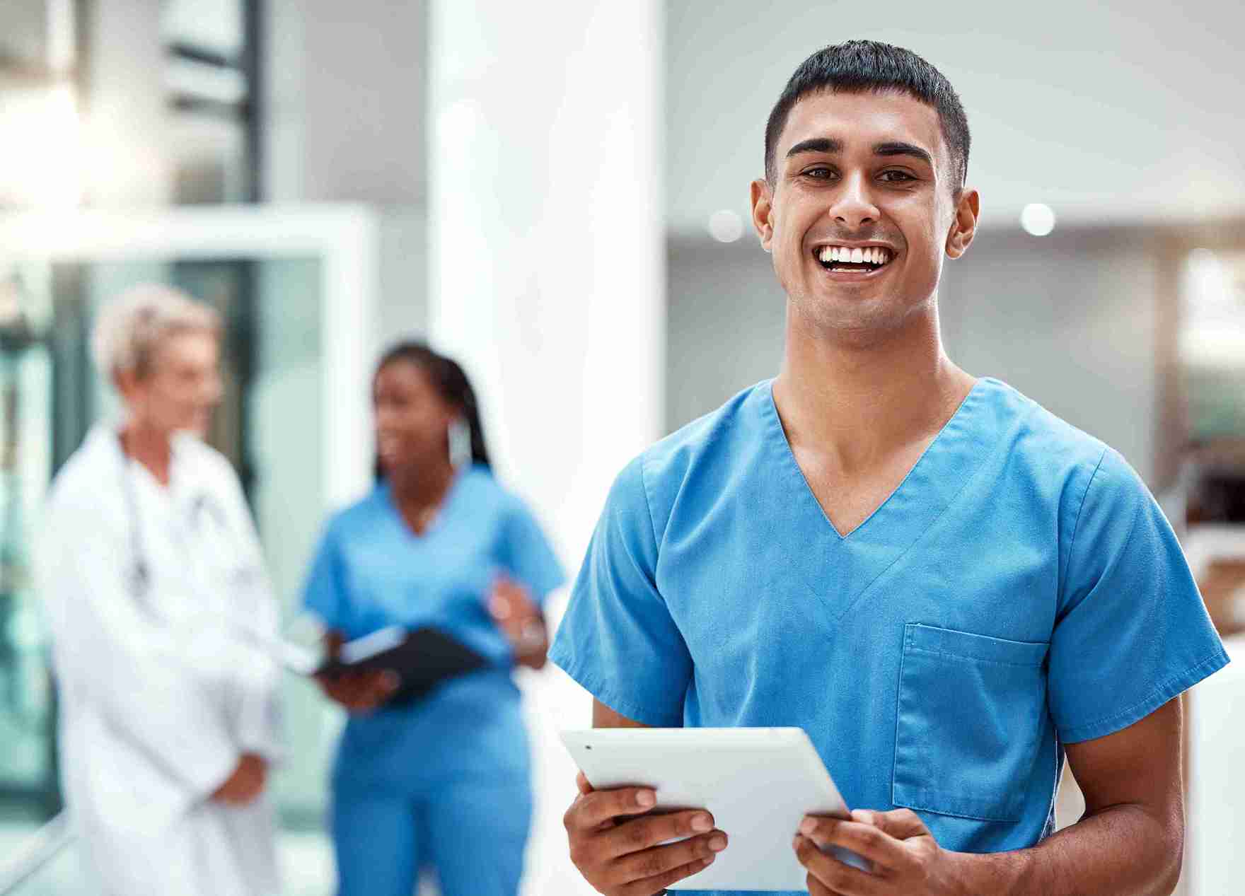A smiling health care worker in blue scrubs stands in a bright medical ...